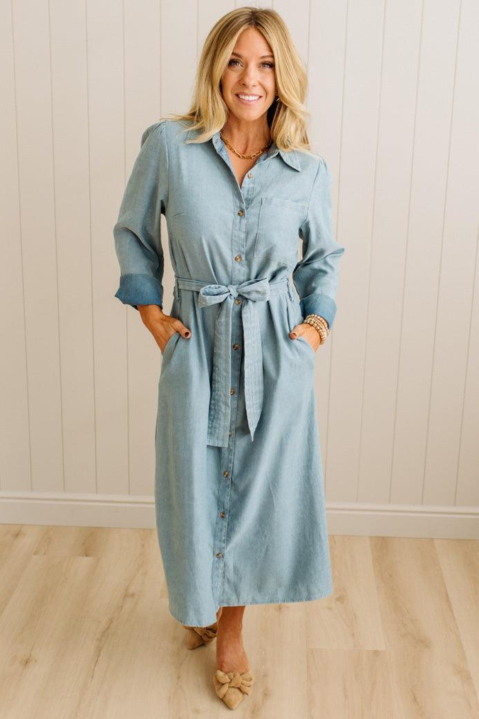 Woman wearing a light blue denim dress with a belt against a white paneled wall.