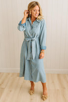 Woman wearing a light blue denim dress with a belt against a white paneled wall.