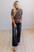 Woman wearing a leopard print top and dark jeans standing in a room with light wooden flooring and white walls.