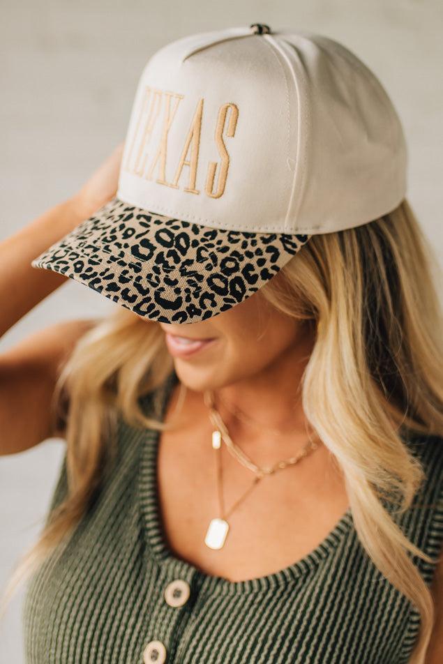 Blonde woman wearing a cream colored trucker hat with leopard print brim, and gold TEXAS embroidered at the front.