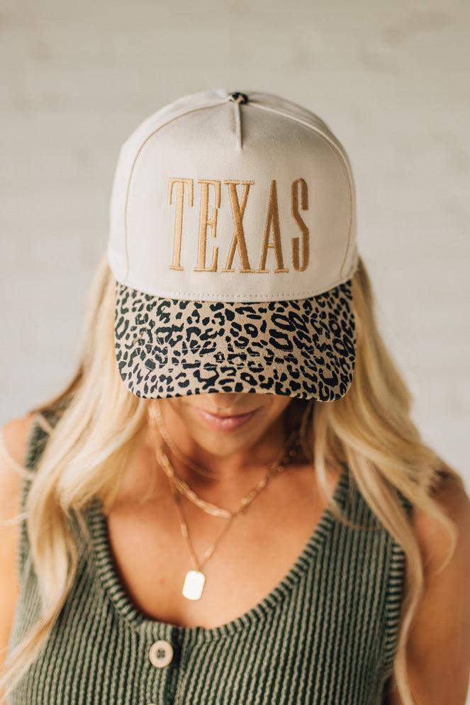 Blonde woman wearing a cream colored trucker hat with leopard print brim, and gold TEXAS embroidered at the front.