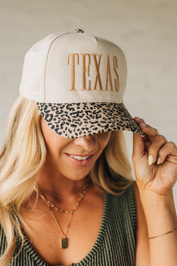 Blonde woman wearing a cream colored trucker hat with leopard print brim, and gold TEXAS embroidered at the front.
