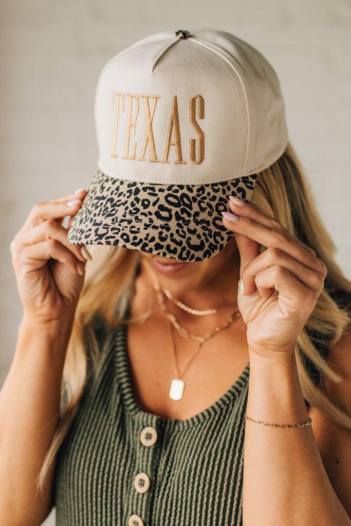 Blonde woman wearing a cream colored trucker hat with leopard print brim, and gold TEXAS embroidered at the front.