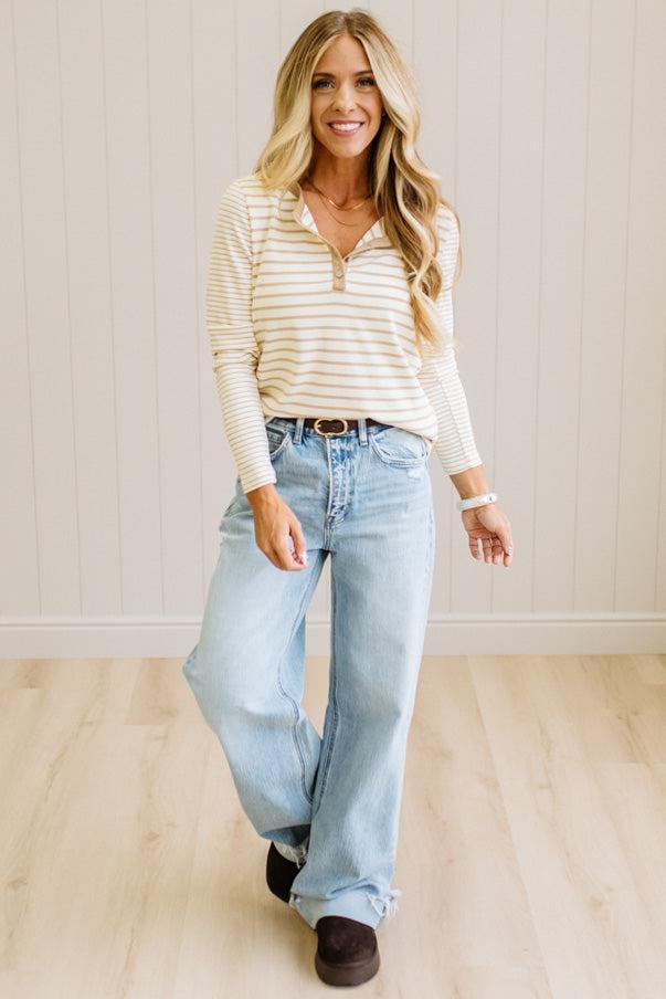 Blonde woman in a brightly lit studio wearing light wide leg jeans, dark clogs, and a striped henley top in cream and taupe.