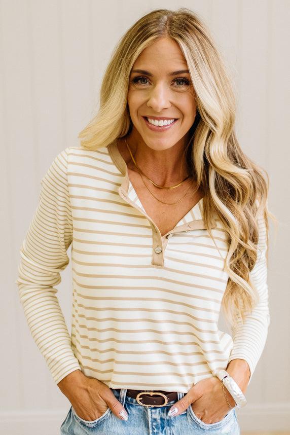 Blonde woman in a brightly lit studio wearing light wide leg jeans, dark clogs, and a striped henley top in cream and taupe.