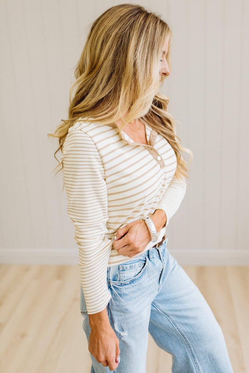 Blonde woman in a brightly lit studio wearing light wide leg jeans, dark clogs, and a striped henley top in cream and taupe.