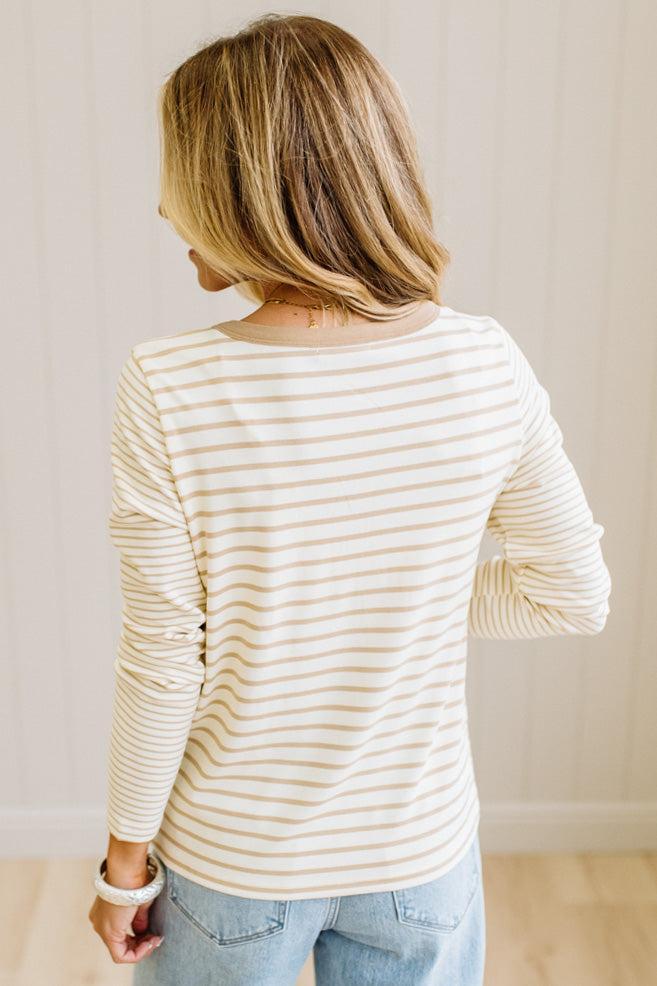 Blonde woman in a brightly lit studio wearing light wide leg jeans, dark clogs, and a striped henley top in cream and taupe.