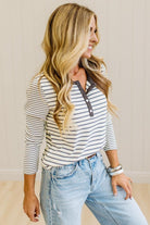 Blonde woman in a brightly lit studio wearing light wide leg jeans, dark clogs, and a striped henley top in cream and charcoal.