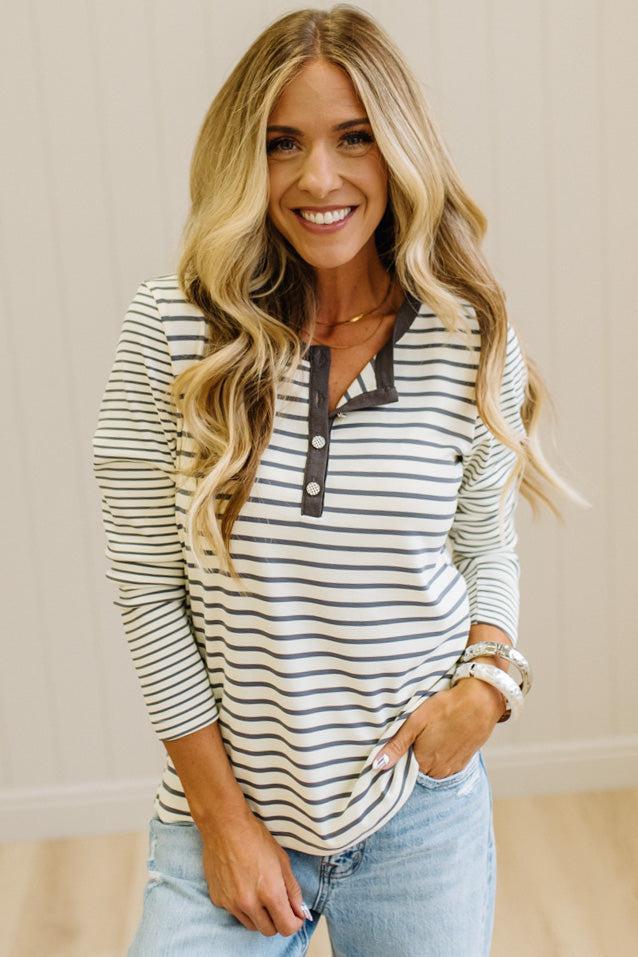 Blonde woman in a brightly lit studio wearing light wide leg jeans, dark clogs, and a striped henley top in cream and charcoal.