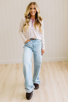 Blonde woman in a brightly lit studio wearing light wide leg jeans, dark clogs, and a striped henley top in cream and lavender.