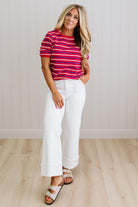 Woman wearing a red and blue striped shirt and white pants standing against a light wooden paneled wall.