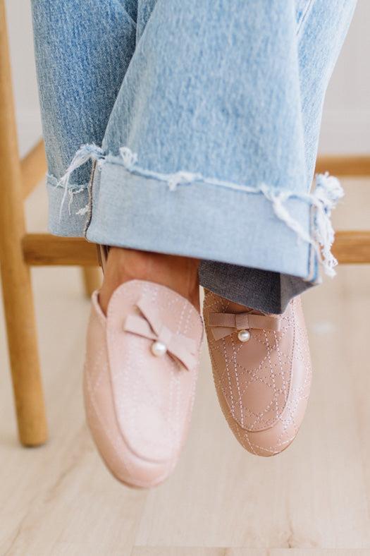 Person wearing pink loafers with pearl embellishments and light blue jeans sitting on a wooden chair.