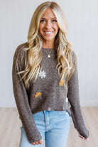 A smiling blonde woman wearing a brown knit sweater with embroidered white and orange flowers, paired with light blue jeans, standing against a light gray paneled wall.