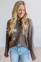 A woman wearing a brown knit sweater with embroidered white and orange flowers, paired with light blue jeans, standing against a white paneled wall.