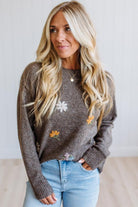 A woman with long blonde hair wearing a brown knit sweater with embroidered white and yellow flowers, paired with light blue jeans, standing against a white paneled wall.