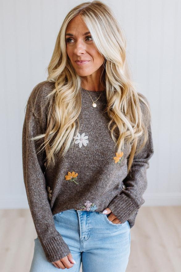 A woman with long blonde hair wearing a brown knit sweater with embroidered white and yellow flowers, paired with light blue jeans, standing against a white paneled wall.