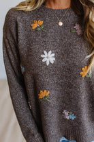 A close-up of a person wearing a dark brown heathered knit sweater with colorful embroidered flowers in yellow, white, pink, and blue. The person is also wearing a delicate gold necklace.