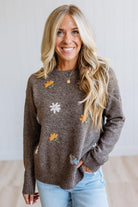 A smiling blonde woman wearing a brown knit sweater with embroidered daisy flowers, paired with light blue jeans, standing against a light-colored wall.