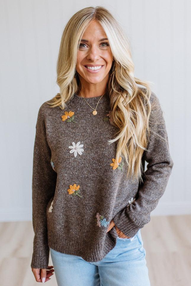 A smiling blonde woman wearing a brown knit sweater with embroidered daisy flowers, paired with light blue jeans, standing against a light-colored wall.