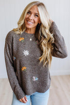 A smiling blonde woman wearing a brown knit sweater with colorful embroidered flowers, paired with light blue jeans, standing against a bright white wall.