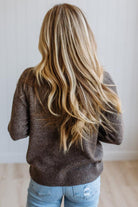 A woman viewed from behind wearing a brown knit sweater and light blue jeans, with long, wavy blonde hair. The background is a clean white paneled wall and light wood floor.