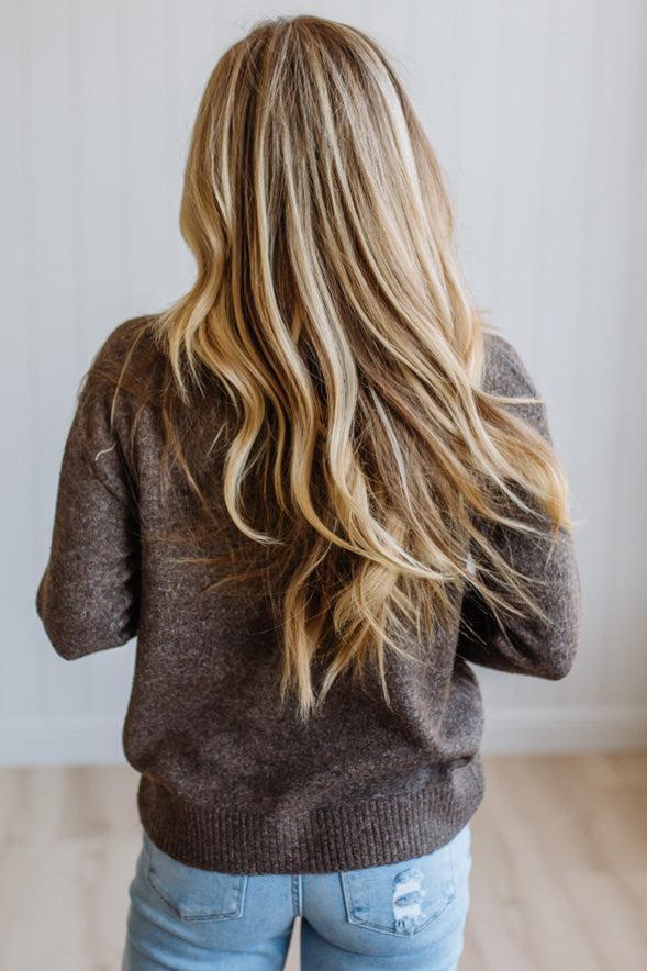 A woman viewed from behind wearing a brown knit sweater and light blue jeans, with long, wavy blonde hair. The background is a clean white paneled wall and light wood floor.