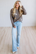 A woman stands in a bright room wearing a brown knit sweater with white and orange floral patterns, light blue flare jeans, and beige pointed shoes.
