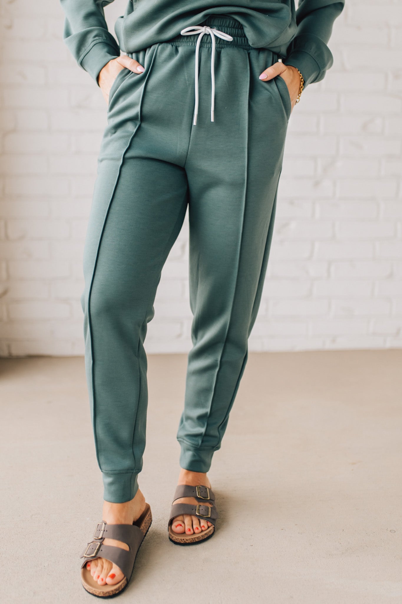 Blonde woman wearing scuba fabric sweatpants with side pockets, a white drawstring on and elastic waistband, and a sewn in center pleat on each leg.