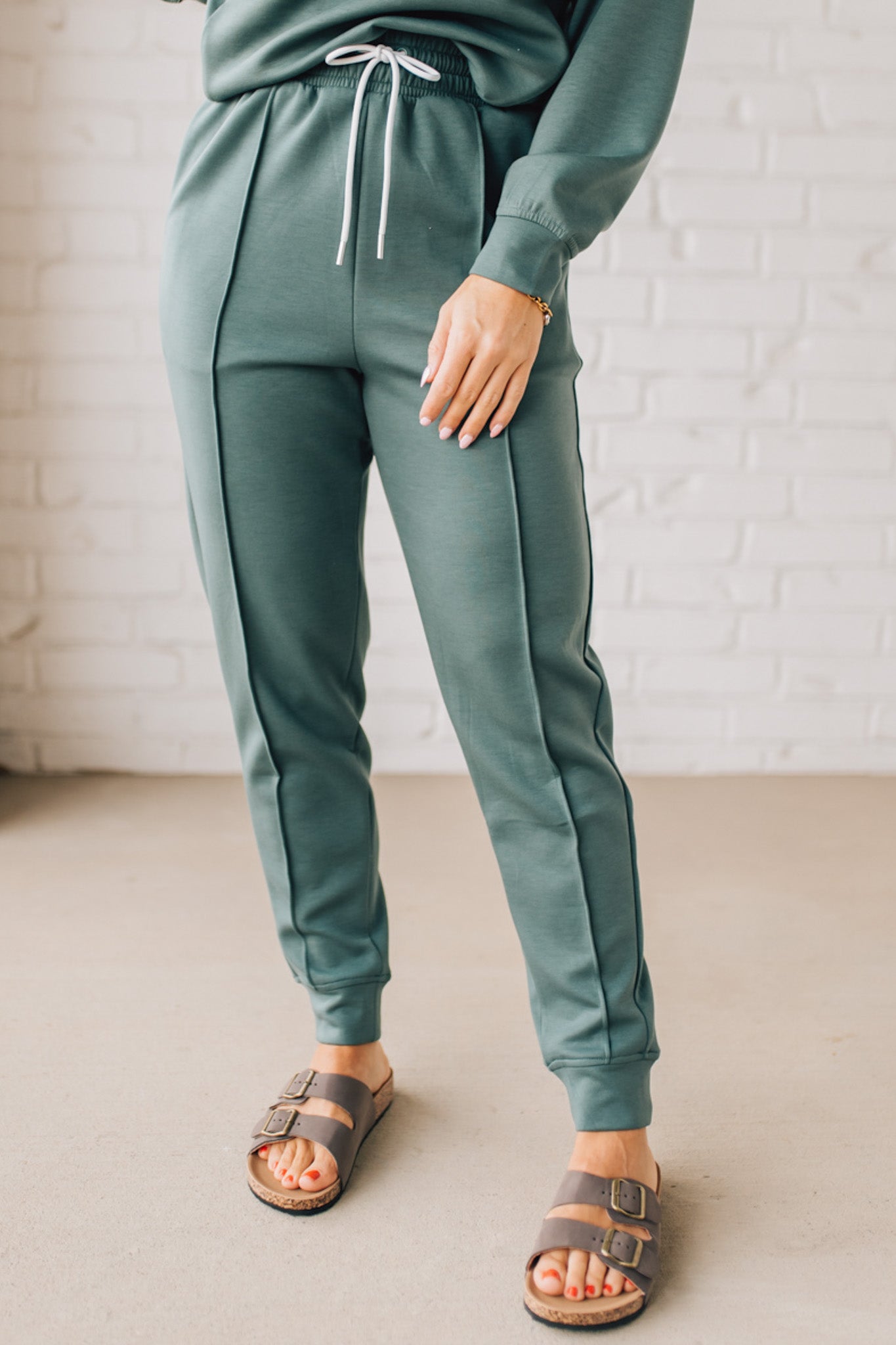 Blonde woman wearing scuba fabric sweatpants with side pockets, a white drawstring on and elastic waistband, and a sewn in center pleat on each leg.