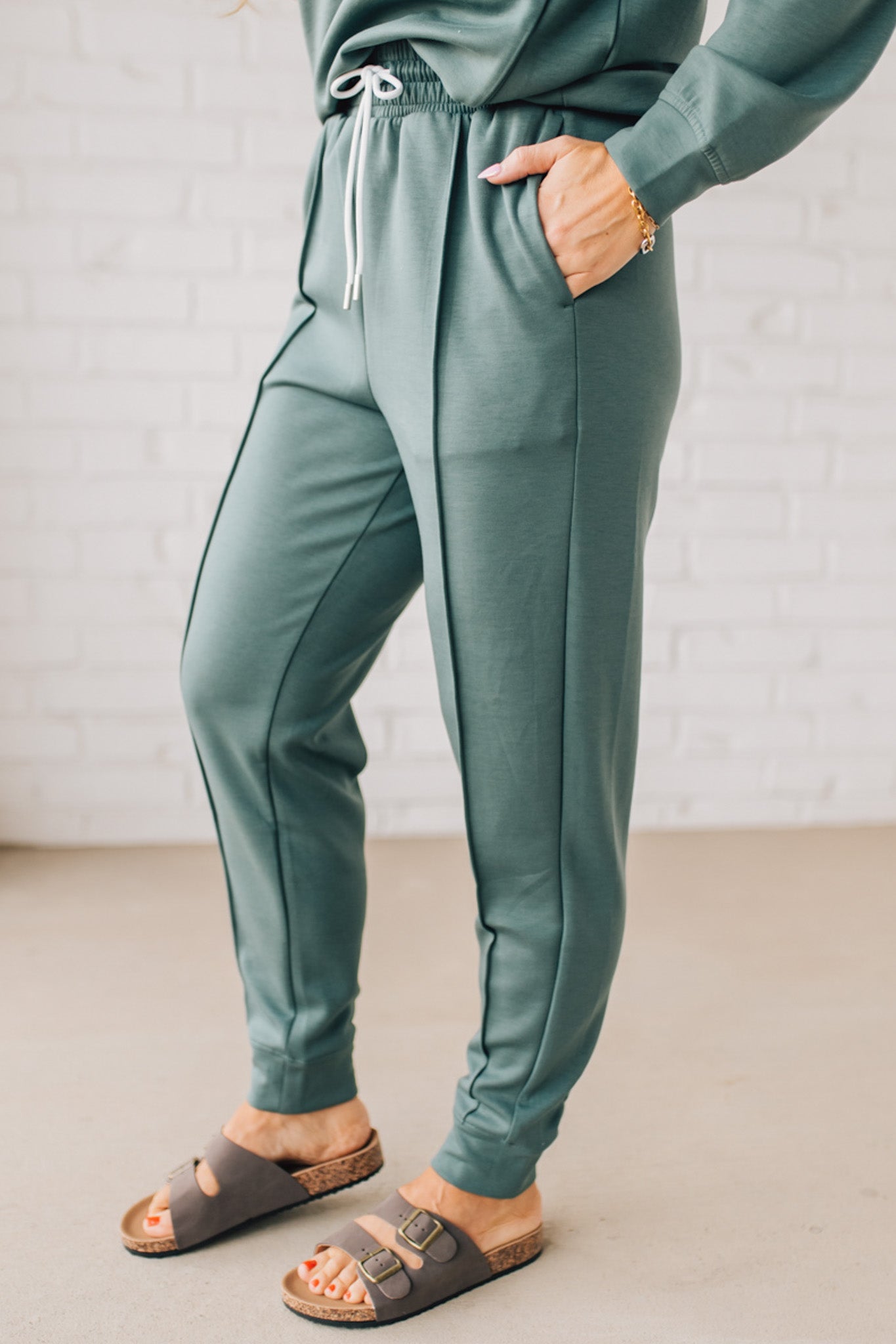 Blonde woman wearing scuba fabric sweatpants with side pockets, a white drawstring on and elastic waistband, and a sewn in center pleat on each leg.