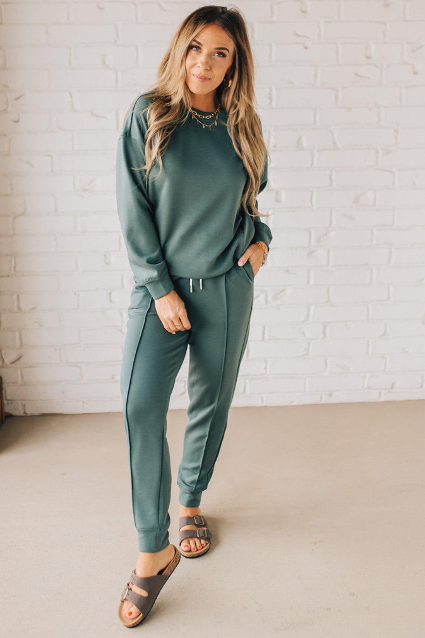 Blonde woman wearing scuba fabric sweatpants with side pockets, a white drawstring on and elastic waistband, and a sewn in center pleat on each leg.