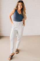 Blonde woman wearing scuba fabric sweatpants with side pockets, a white drawstring on and elastic waistband, and a sewn in center pleat on each leg.