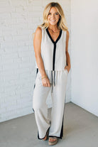 Woman wearing a white sleeveless top and pants with black trim against a white brick wall.
