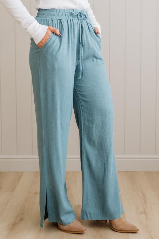 Person wearing teal pants with a white shirt and brown shoes on a light wooden floor.