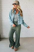 Woman wearing a denim jacket, gray top, green pants, and beige cap against a white brick wall.