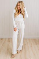 Woman wearing a white outfit standing against a light wooden paneled wall.