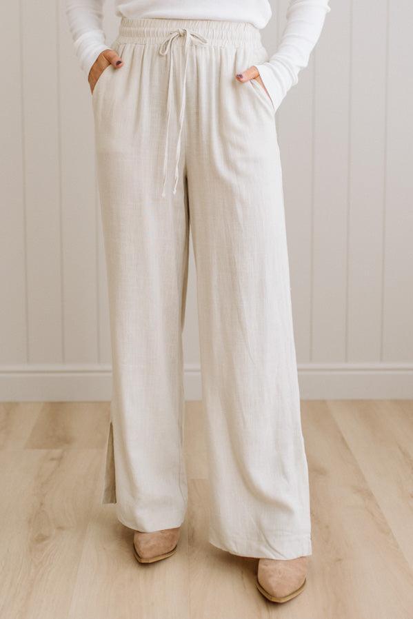 Person wearing beige wide-leg pants and a white top against a light wooden floor and wall background.