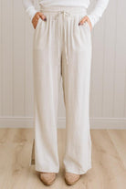 Person wearing beige wide-leg pants and a white top against a light wooden floor and wall background.