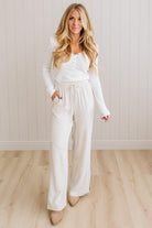 Woman wearing a white outfit standing against a light wooden paneled wall.