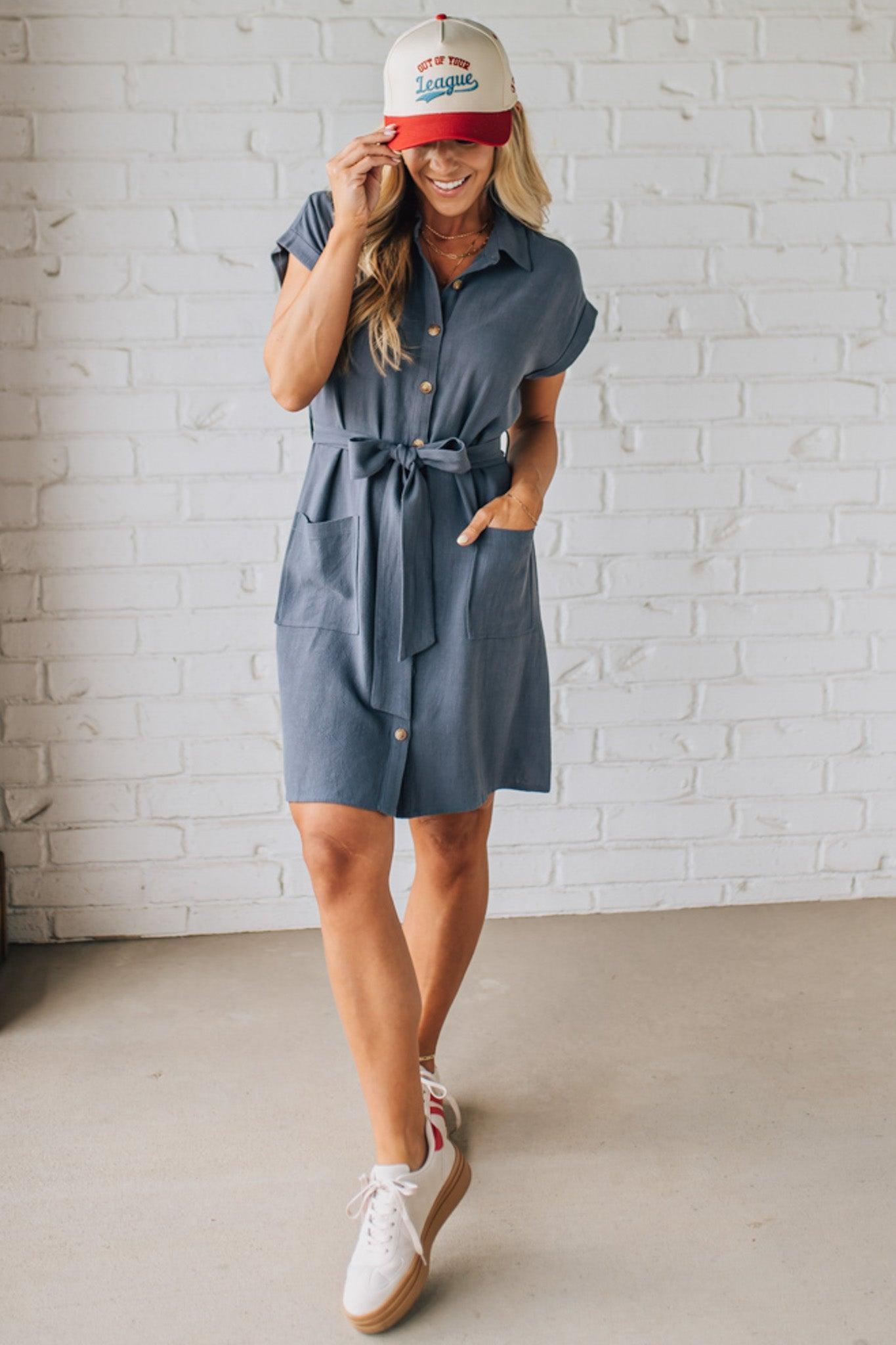 BLONDE WOMAN WARING A KNEE LENGTH SHORT DRESS WITH  BUTTON UP AND COLLARED FRONT. HAS SHORT SLEEVES, FRONT PATCH POCKETS AND TIES AT THE  WAIST, ALL IN SLATE BLUE WITH A TEXTURED LINEN BLEND.