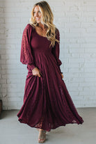 Woman wearing a burgundy dress with lace details against a white brick wall.