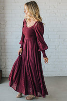 Woman wearing a burgundy dress with long sleeves against a white brick wall.