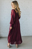 Woman wearing a burgundy dress with lace details against a white brick wall.