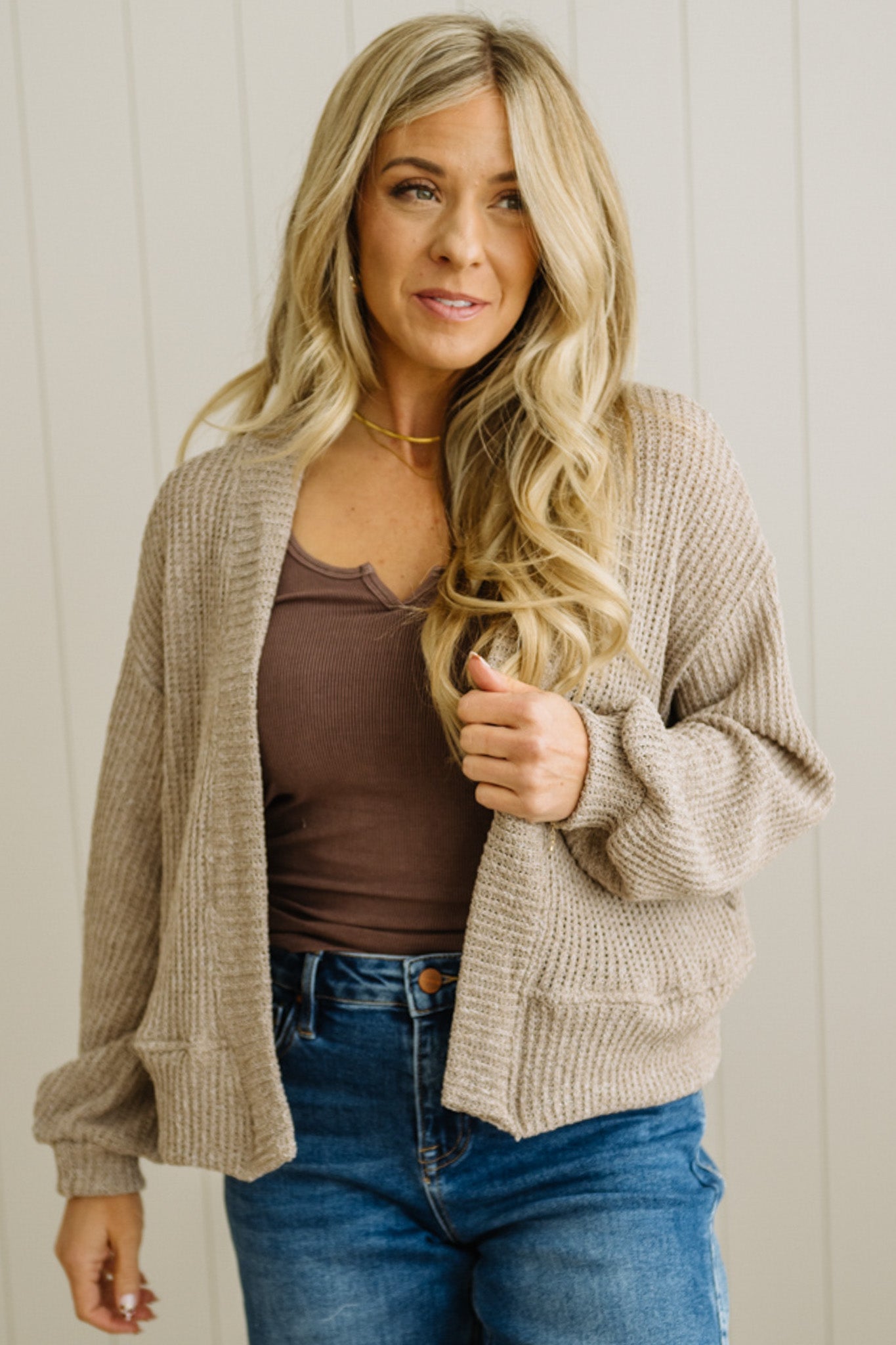 Woman wearing a beige cardigan over a brown top and blue jeans against a neutral background