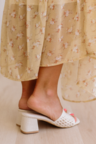 White open-toe sandals worn with a floral dress on a light wooden floor.