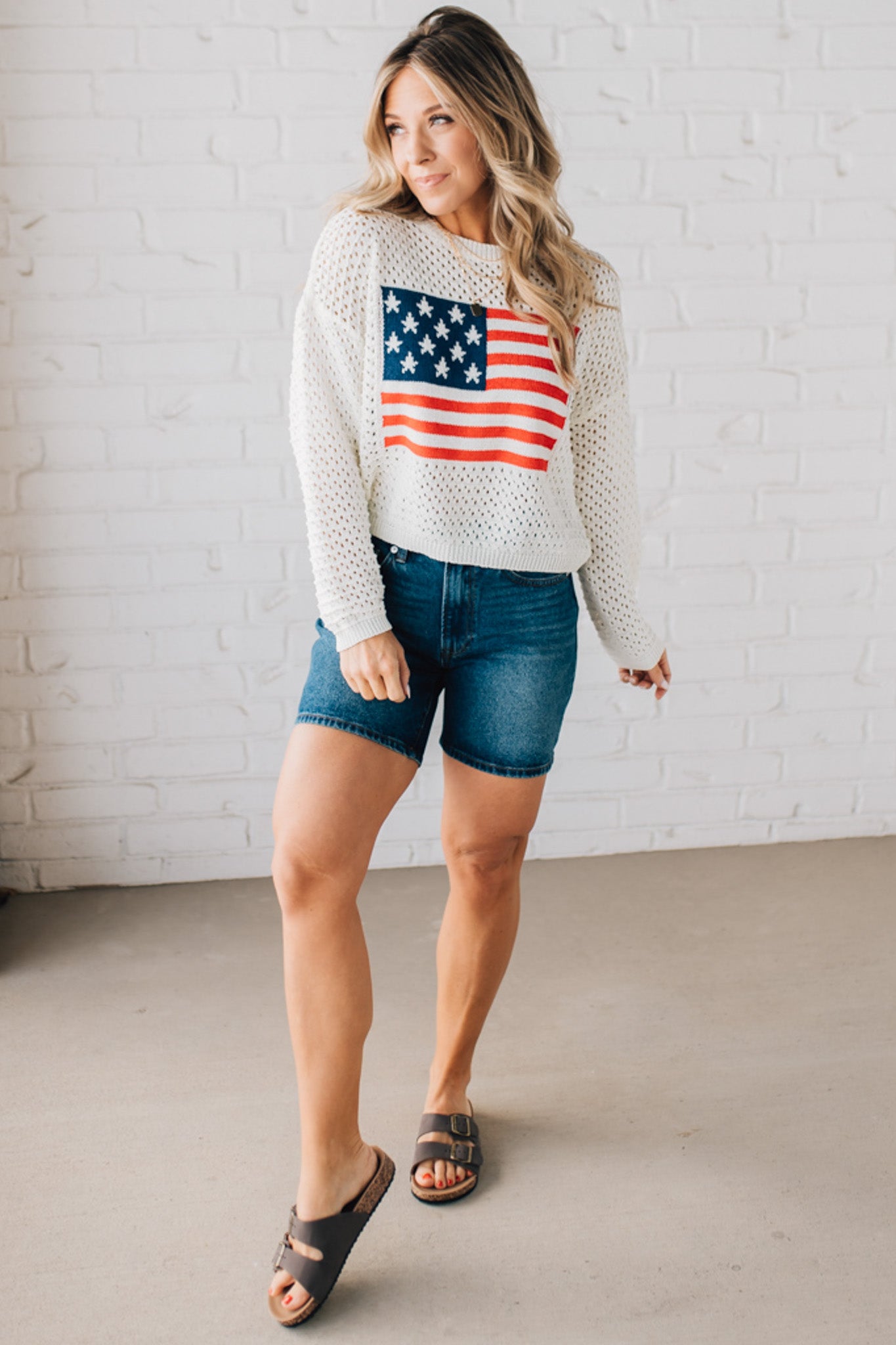 Blonde woman wearing a loose knit low gauze white sweater with drop shoulder and woven American flag at center front.