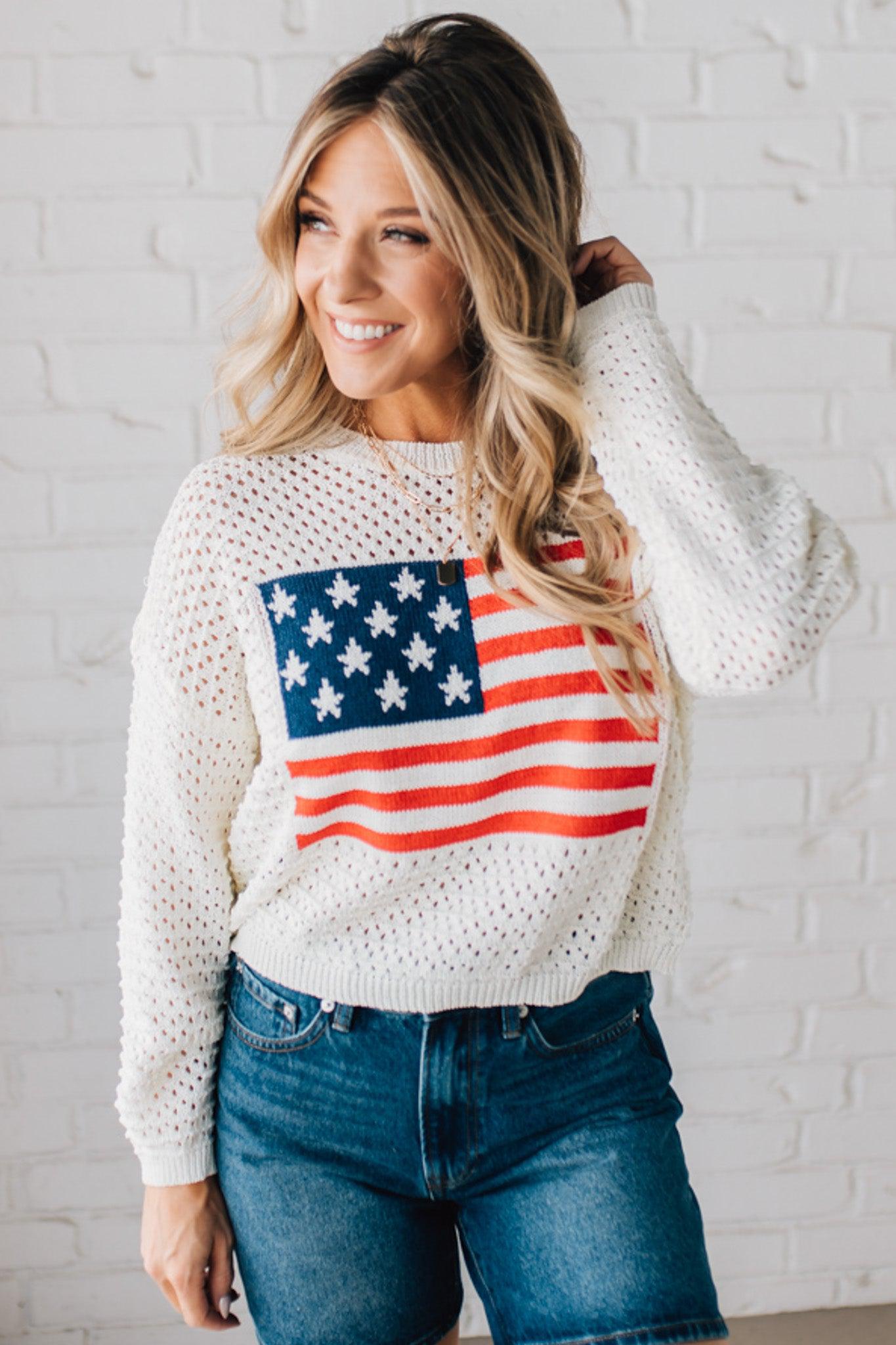 Blonde woman wearing a loose knit low gauze white sweater with drop shoulder and woven American flag at center front.