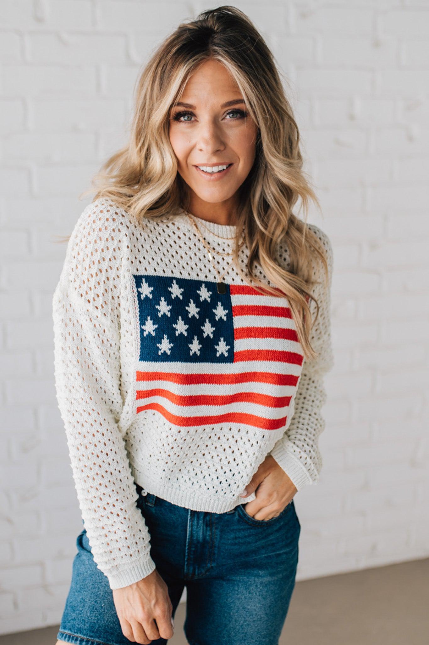 Blonde woman wearing a loose knit low gauze white sweater with drop shoulder and woven American flag at center front.