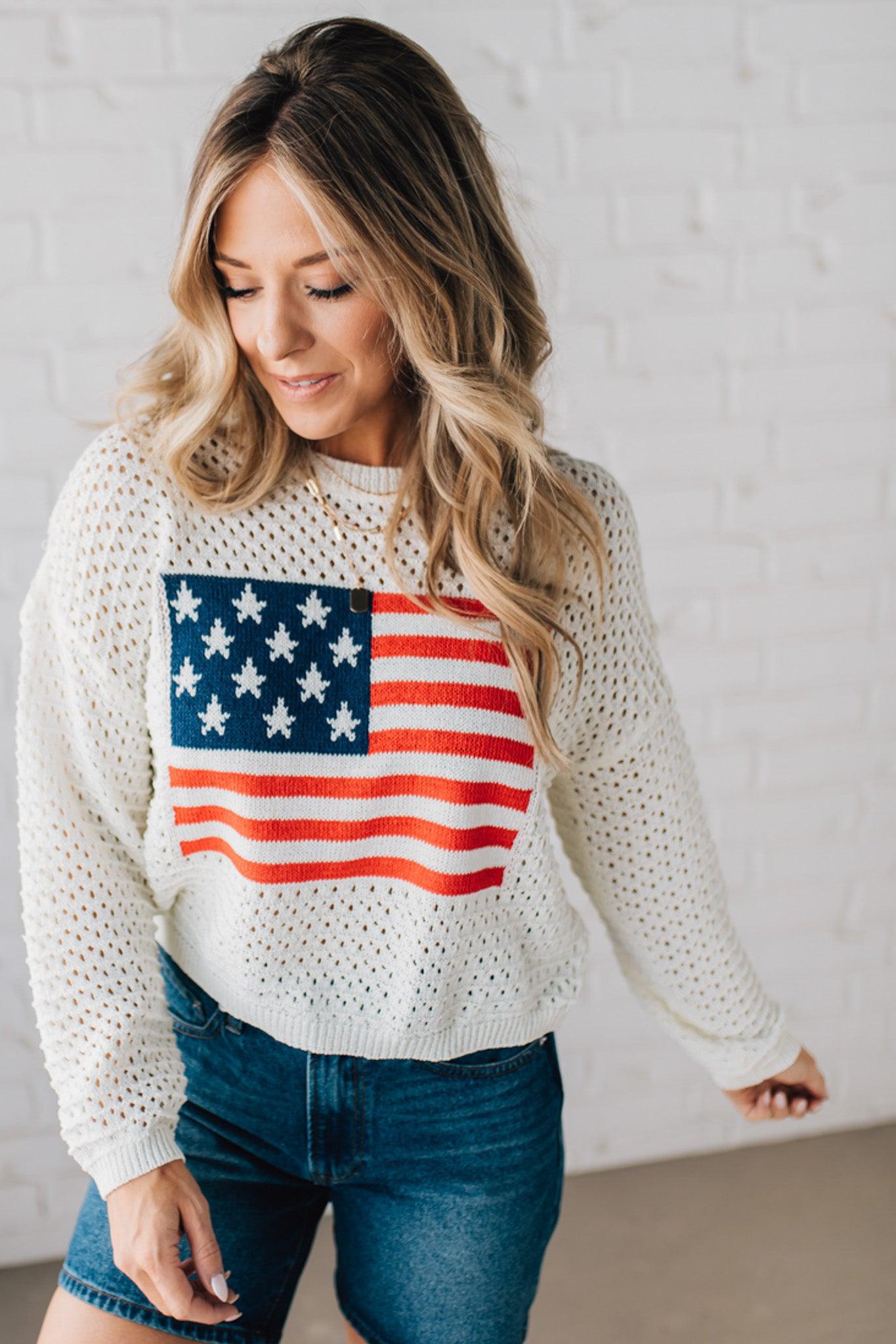 Blonde woman wearing a loose knit low gauze white sweater with drop shoulder and woven American flag at center front.