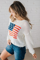 Blonde woman wearing a loose knit low gauze white sweater with drop shoulder and woven American flag at center front.